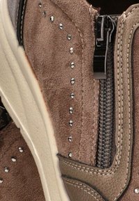 Detalle de zapato de ante marrón con tachuelas metálicas, costura beige, cremallera lateral y borde de suela texturizada.