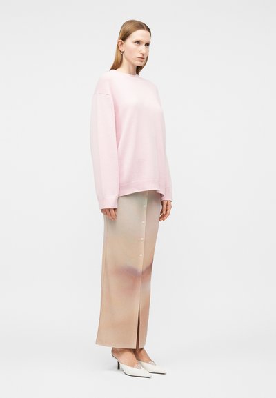 Γυναίκα στέκεται προς τα αριστερά φορώντας ένα φούξια ανοιχτό ροζ φαρδύ πουλόβερ, μια μακριά μπεζ φούστα με κουμπιά και λευκά μυτερά τακούνια σε απλό φόντο.