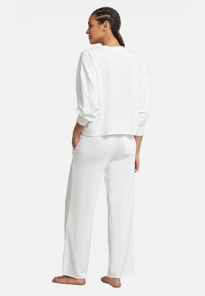 Γυναίκα με πλεξούδες, φοράει λευκό, φαρδύ μακρυμάνικο τοπ και φαρδιά παντελόνια, στέκεται ξυπόλητη με τα χέρια στις τσέπες, γυρίζοντας την πλάτη.