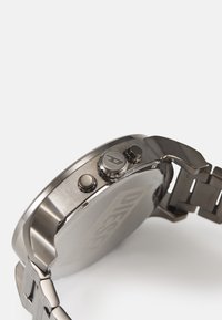 Montre en acier inoxydable avec un boîtier rond à finition brossée. Elle est équipée de deux boutons poussoirs sur le côté et d'un bracelet en métal maillé.