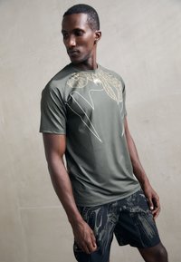T-shirt de sport vert olive avec un motif graphique doré sur la poitrine, associé à un short noir présentant des designs et textures abstraits.