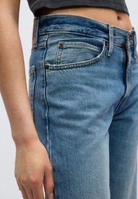 Κοντινό πλάνο ατόμου που φορά ψηλόμεσα, ανοιχτού μπλε τζιν και κοντό σκούρο τοπ, εμφανίζοντας τη μέση, τις τσέπες και το αριστερό χέρι.