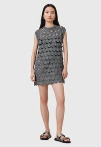 Rochie gri fără mâneci, cu un model texturat sub formă de cercuri, tiv ondulat și croi lejer. Modelul poartă sandale negre cu accesorii decorative.