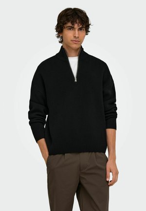 Jeune homme portant un pull noir à demi-fermeture éclair par-dessus une chemise blanche et un pantalon marron, debout avec une main dans la poche sur un fond uni.