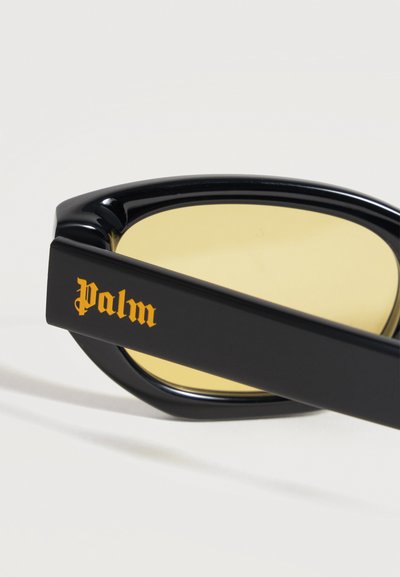 Branché de lunettes de soleil noir avec verre teinté jaune et "Palm" écrit en police gothique jaune sur le côté.