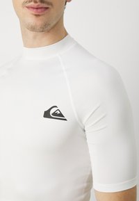 Hvit kortermet rash guard med rund hals og svart logoemblem på brystet. Glatt, stretchy stoff med flate sømmer.