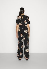 Jumpsuit floral con fondo negro, que presenta mangas cortas, una cintura ajustada y pantalones de pierna ancha con patrones de flores rosas y blancas.