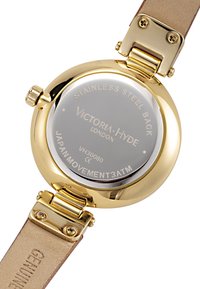 Montre en acier inoxydable doré au dos gravé de détails. Équipée d'un bracelet en cuir tan et d'accents en métal poli. Résistante à l'eau jusqu'à 3 ATM.