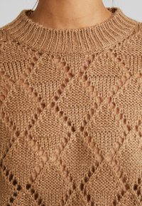 Gros plan d'une personne portant un pull en tricot marron clair avec un motif ajouré en forme de losange et un col rond côtelé.