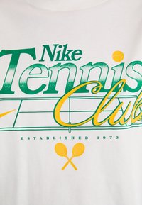 Valge puuvillane t-särk, millel on roheline ja kollane graafikatekst "Nike Tennis Club" tennisereketi disaini kohal, asutatud 1972.