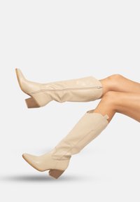 Botas de cowboy de cuero beige con puntera puntiaguda, tacón bloque de altura media, caña caida y cierre de cremallera lateral. Textura suave con detalles curvados.