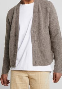 Ljusbrun stickad kofta med ribbad textur, öppen front, fem knappar och långa ärmar, bärs över en vit skjorta och beige kordflordingbyxor.
