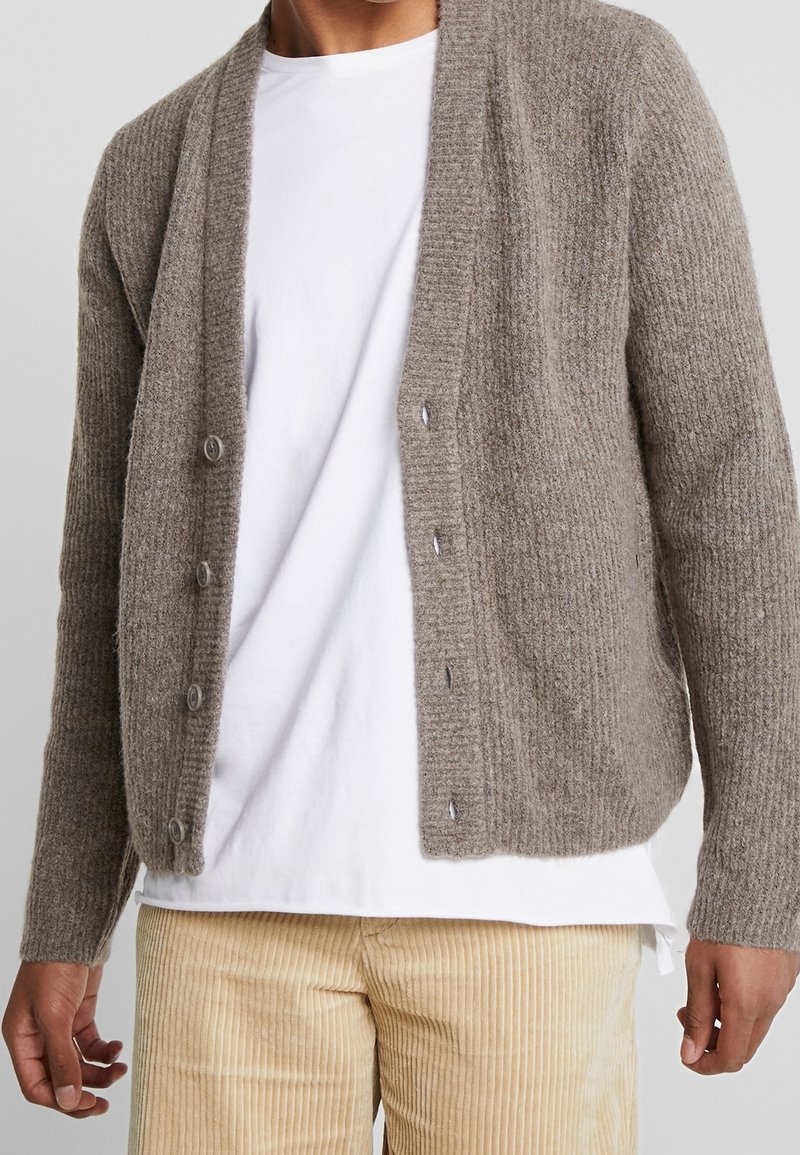 Ljusbrun stickad kofta med ribbad textur, öppen front, fem knappar och långa ärmar, bärs över en vit skjorta och beige kordflordingbyxor.