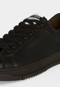 Μαύρο sneaker από υφασμένο δέρμα με μαύρα κορδόνια, καφέ ρουθούνια, σκούρα σόλα και λευκή ετικέτα στη γλώσσα με μερικό κείμενο της μάρκας.