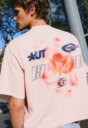 Mladý muž nosí bledo ružové tričko s veľkou potlačenou oranžovou ružou, čiernym textom a grafickými návrhmi na chrbte, vonku za denného svetla.