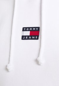 Sudadera blanca con cordones ajustables, que presenta un parche bordado rectangular con "TOMMY JEANS" y acentos en rojo, blanco y azul.