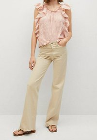 Vaaleanpunainen hihaton toppi, jossa on röyhelöillä varustetut olkapäät, yhdistettynä beigekirjaviin löysiin farkkuihin. Malli seisoo toisella kädellään lantiolla, jalassaan ruskeat varvastossut.