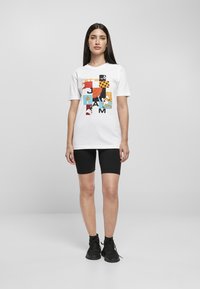 Hvit t-skjorte med et fargerikt grafisk design med teksten "LET'S JAM" og tegneseriefigurer. Kombinert med svarte sykkelhorts og svarte sko.