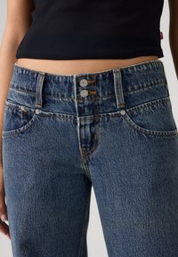 Μπλε τζιν παντελόνι με ψηλή μέση, δύο ασημένια κουμπιά και αντιθετική πορτοκαλί ραφή. Διαθέτει μπροστινές τσέπες και υφασμάτινη υφή.