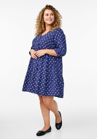 Marineblå kjole med hvide prikker, V-udskæring, knapdetalje, trekvartærmer, flagrende nederdel og en tiered bundruffle.