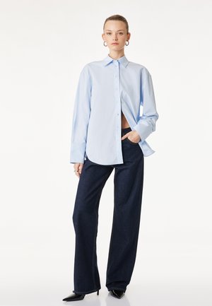 Femme en chemise oversize bleu clair boutonnée, jean large foncé, talons pointus noirs, main dans la poche, debout devant un fond blanc.