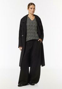 Schwarzer langer Mantel, gemusterte schwarz-weiße Bluse und weite schwarze Hose. Glatte Texturen mit Oversized-Passform. Runde Knopfdetails am Mantel.