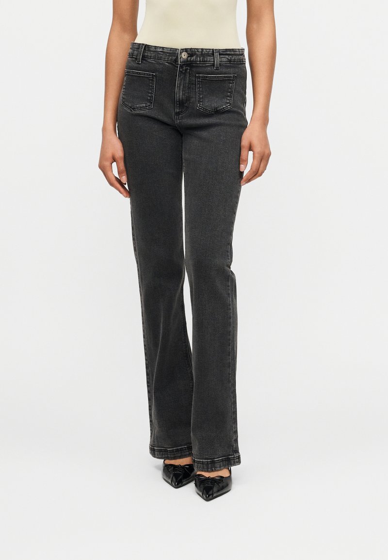 Femme portant un jean noir taille haute à jambes droites et des talons noirs à bout pointu, debout devant un fond blanc uni.