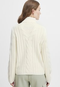 Pull en tricot de couleur crème avec un col haut, présentant des côtes verticales et un motif en torsades au dos et sur les manches.