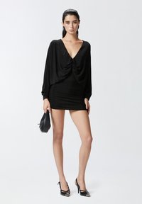 Mini abito nero con corpino drappeggiato, scollatura a V profonda, maniche lunghe e dettagli arricciati. Abbinato a décolleté slingback a punta e una piccola borsa.