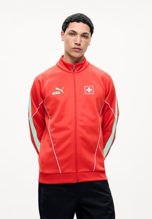 Jeune homme portant une veste de sport rouge zippée avec des liserés blancs, un emblème du drapeau suisse et le logo Puma avec le texte « KING », debout les mains derrière le dos.