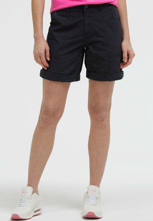 Frau trägt schwarze, umgeschlagene Shorts, pinkes Oberteil und weiße Sportschuhe mit pinker Sohle, steht vor weißem Hintergrund.