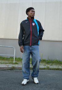 Sort vindjakke med blå detaljer og rød lynlås, med et Atletico Madrid-logo, parret med løse blå jeans og hvide sportsko.