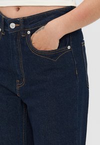 Mørkeblå denimjeans med høy midje, med kobbertråd, frontlommer og en glatt tekstur. Standard knappelukking.
