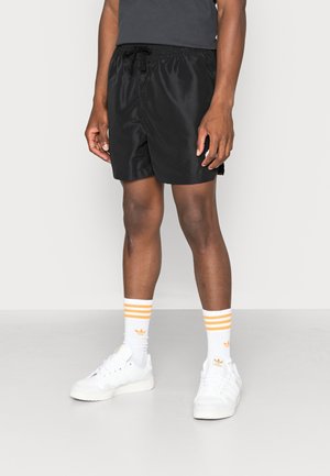 Man draagt zwarte shorts, witte sneakers en witte sokken met drie oranje strepen, staand tegen een effen lichte achtergrond.
