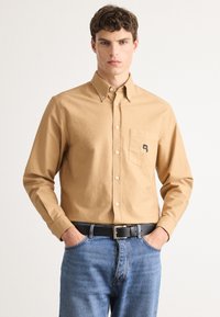 Ungdomsmann i en beige skjorte med knapp foran og brystlomme med logo, blå jeans og svart belte mot en hvit bakgrunn.