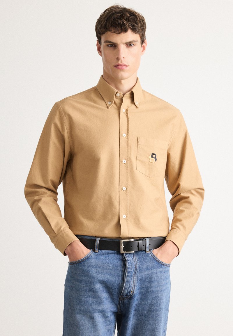 Ungdomsmann i en beige skjorte med knapp foran og brystlomme med logo, blå jeans og svart belte mot en hvit bakgrunn.