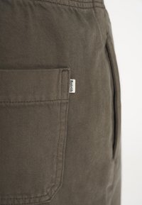 Brune bomuldsbukser med en sidelomme og et lille mærke. Stoffet har en subtil tekstur med en solid, dæmpet farveskala.