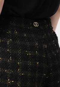 Juodos ir auksinės langelių rašto tweed kelnės su tekstūruotu paviršiumi, kuris puošia aukso spalvos apvaliu mygtuku liemens srityje.