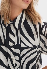 Blusa nera e bianca a maniche corte con motivi astratti a foglia, colletto classico e bottoni, realizzata in un tessuto leggero e testurizzato.