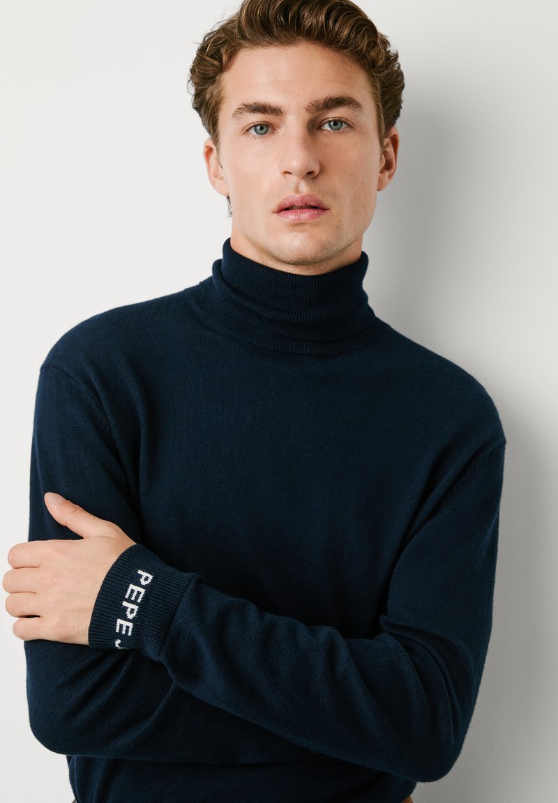 Granatowy sweter typu golf wykonan z miękkiego materiału, z prążkowanym dekoltem i mankietami oraz białym napisem "PEPE". Dopasowany krój, minimalistyczny styl.