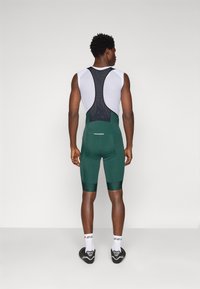 Shorts de vélo pour hommes en vert foncé, avec un dos en mesh, une coupe ajustée et des accents noirs. Portés avec des chaussettes blanches et des chaussures de cyclisme.
