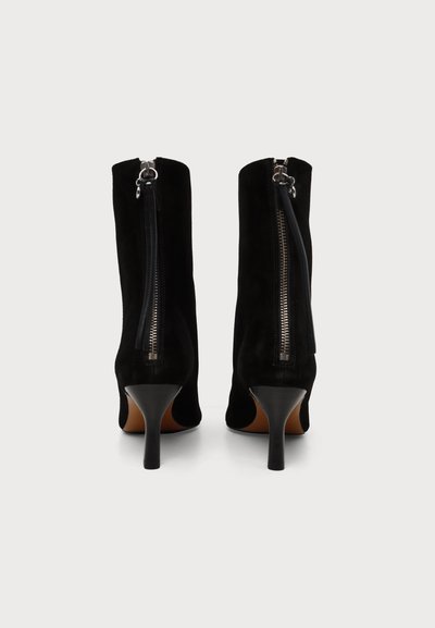 Paire de bottines en daim noir avec talons hauts et fins et fermetures éclair verticales à l'arrière sur fond blanc.