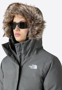 Grå isoleret jakke med hætte i faux pels, der har lynlås foran, trykknapper og The North Face-logo i hvid.