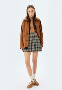 Bruine suède jas over een beige trui, gecombineerd met een geruite geplooide rok en bruine sneakers; witte crew sokken zichtbaar.