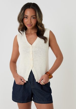 Frau mit welligem Haar trägt eine weiße ärmellose Strickweste mit Knöpfen und dunkle Jeansshorts, accessoriert mit Ringen und einem durchsichtigen Armband.