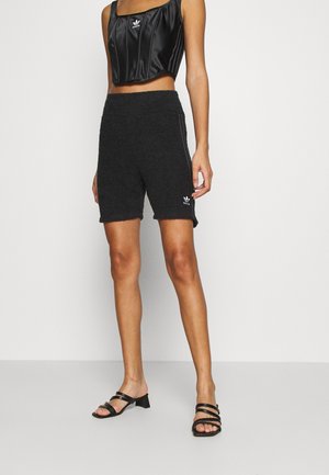 Vrouw draagt zwarte gestructureerde Adidas short, zwarte leren Adidas crop top en zwarte sandalen met hak, staand tegen een witte achtergrond.