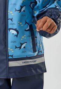 Enfant portant un imperméable bleu à motifs de requins et de poissons, poignets bleu marine, main visible et logo "Swedemount" sur une bande réfléchissante.