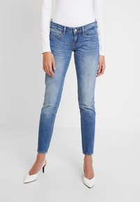 Mujer vistiendo jeans ajustados azul y una blusa blanca de manga larga, combinada con tacones blancos de punta, de pie contra un fondo simple.