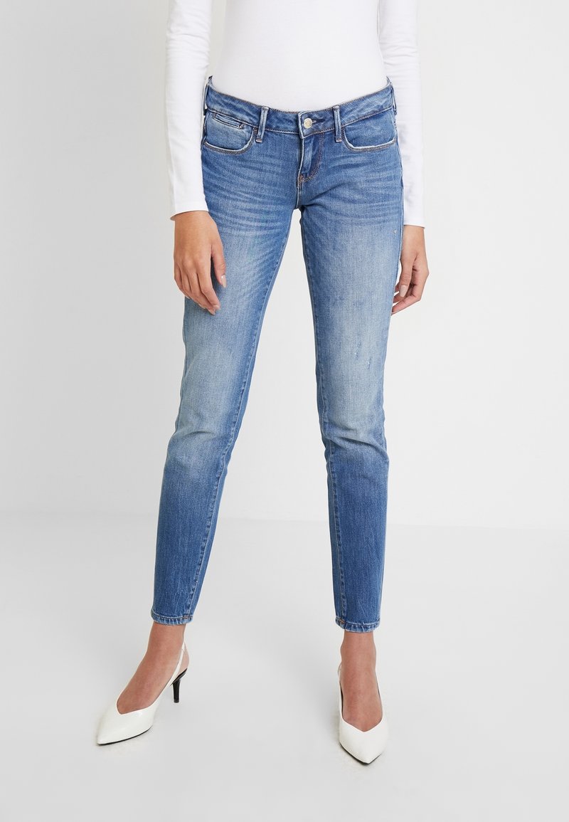 Mujer vistiendo jeans ajustados azul y una blusa blanca de manga larga, combinada con tacones blancos de punta, de pie contra un fondo simple.