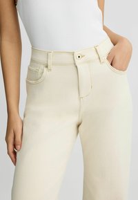 Jean haut de taille en denim couleur crème, avec des poches avant, une fermeture avec bouton en haut et de subtils détails de couture le long des coutures.